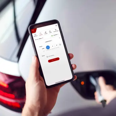 Smartfox-App auf dem Smartphone steuert eine angebundene Ladestation, im Hintergrund lädt ein Elektroauto